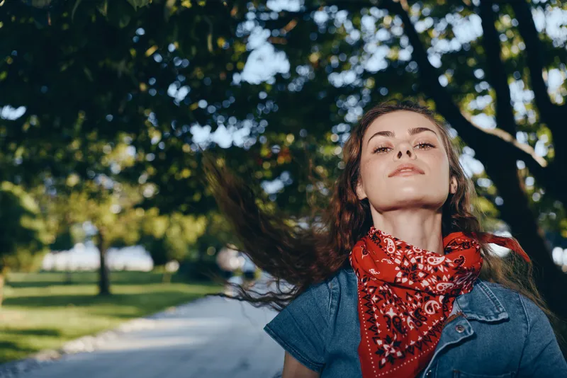 young woman outdoors with red bandana and flowing hair enjoying sunny day in green park with tall trees and pathway in background