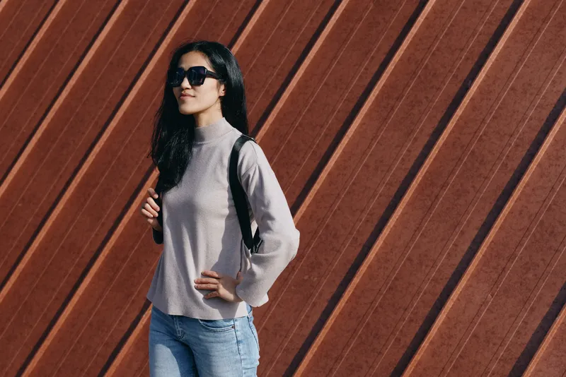 Woman sunglasses sweater backpack jeans outdoor fashion style casual pose confidence against a wooden wall, a stylish, modern look suited for streetwear and daytime shoots