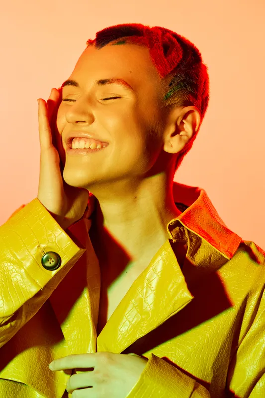 Smiling woman with colorful hairstyle and yellow jacket enjoying vibrant studio lighting, expressing happiness and confidence with closed eyes and radiant smile.