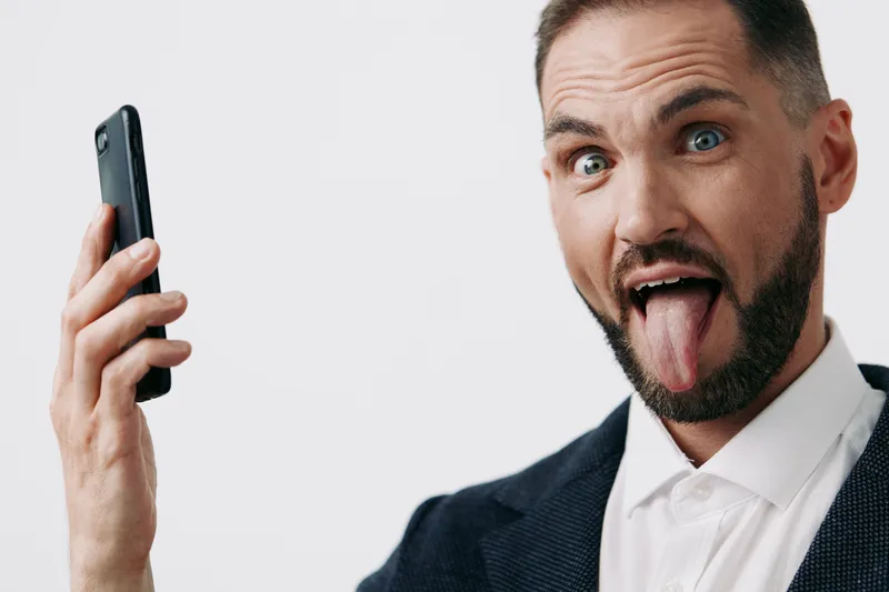 A professional businessman against an isolated colored background, showing playful emotion as he holds a smartphone. The scene conveys confidence, curiosity, and modern corporate energy.