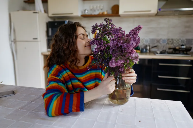 A woman in a colorful striped sweater admires a bouquet of purple flowers on a kitchen table, creating a cozy home moment focused on fragrance and simple daily joy.