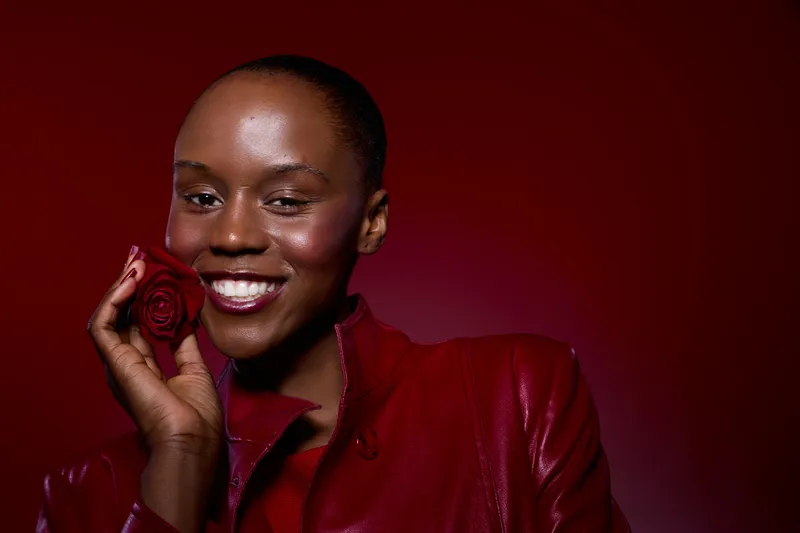 Smiling African woman with dark skin holds red rose near face wearing red leather jacket. Studio portrait on red background. Concept of beauty, fashion, elegance, and confidence.