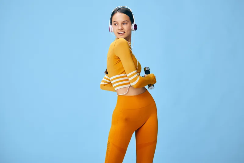 Fitness, sport woman in orange athletic wear posing with a dumbbell against a blue studio backdrop, headphones on, showing strength, balance and confident workout energy.