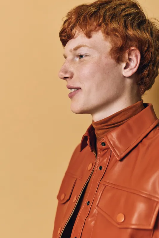 Profile of young man with red hair wearing orange jacket against beige background. Casual style with natural expression and side view of face for fashion or portrait concept.