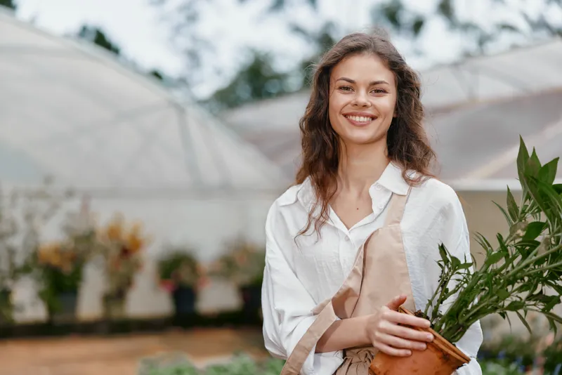 Smiling young woman in a greenhouse holding plants, wearing a white shirt and apron, showcasing a joyful gardening concept with vibrant greenery