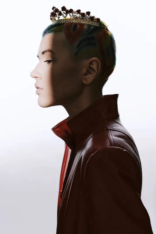 Profile of a stylish woman with short colorful hair and floral crown, wearing a red leather jacket, showcasing unique fashion and creative expression in minimalist lighting.