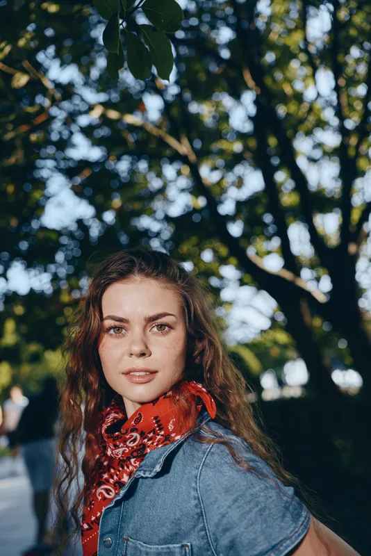 smiling woman with red scarf and denim jacket standing outdoors under lush green trees in soft natural light, warm atmosphere, casual style, happy expression
