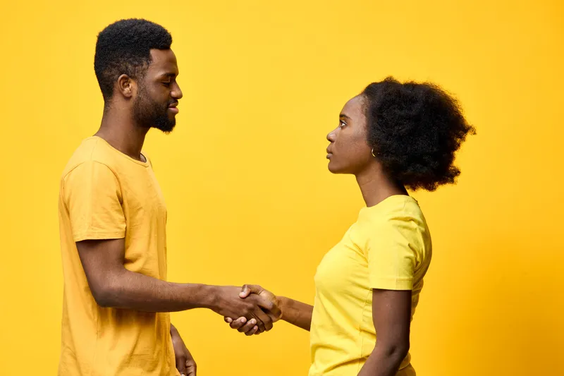 Two young African individuals shaking hands with smiles on a bright yellow background, representing friendship, unity, and collaboration in a modern professional setting