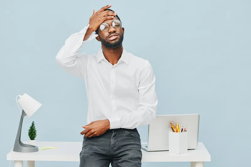 Frustrated young man with glasses in white shirt standing at desk with laptop, pen holder and desk lamp on light blue background. People lifestyle concept, workplace stress, office problem, stressful 