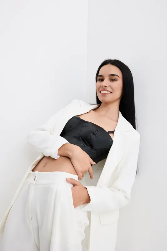 Smiling woman in white suit with black crop top posing relaxed against white wall, showcasing modern fashion and confident style with minimalistic background.