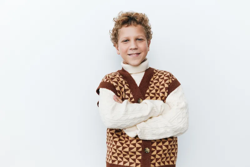 Smiling young boy stands with arms crossed, wearing a chunky geometric knit vest over a turtleneck, against a clean white studio backdrop.