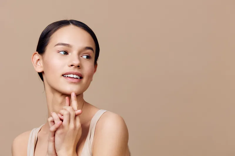 Young woman with a joyful expression, wearing a light outfit, against a beige background, showcasing natural beauty and healthy skincare