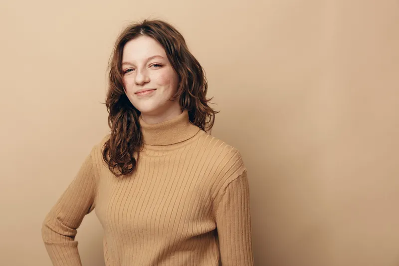 Young woman with curly brown hair wearing beige turtleneck sweater smiles confidently against plain beige background. Concept of casual style, natural beauty, and positive mood.