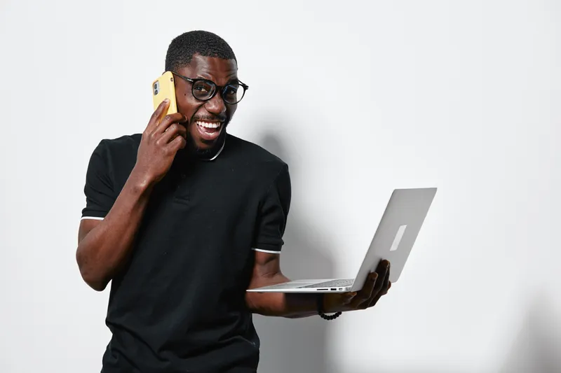 Black man with dark skin talking on smartphone and holding laptop in hand, smiling and standing against white background for technology and communication concept.