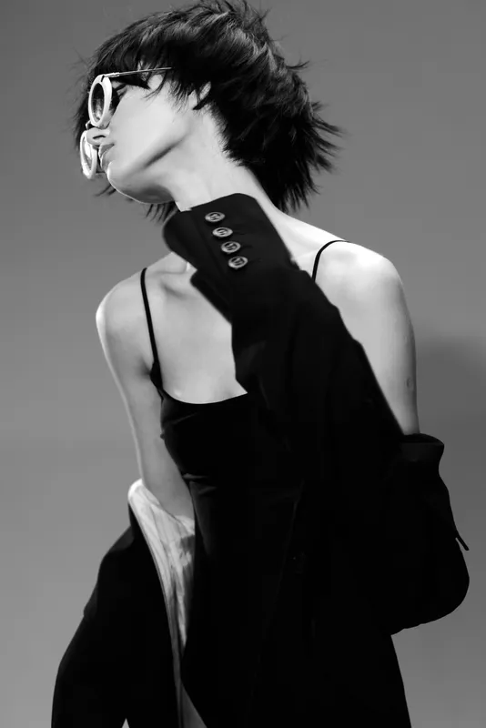 Black and white portrait of a stylish woman wearing sunglasses and a blazer, expressing elegance and modern fashion in a minimalist setting.