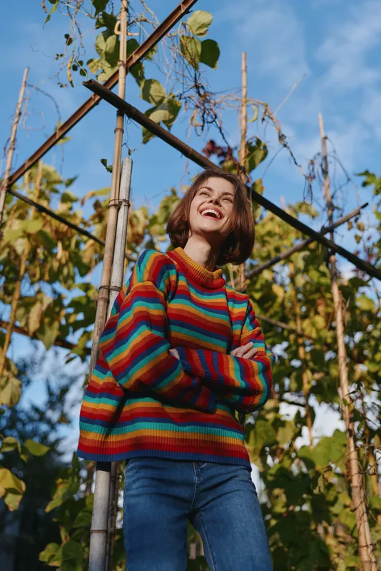 Young woman in her early 20s wearing a colorful striped sweater and jeans, smiling woman with crossed arms in a sunny outdoor garden by trellis plants, joyful lifestyle.