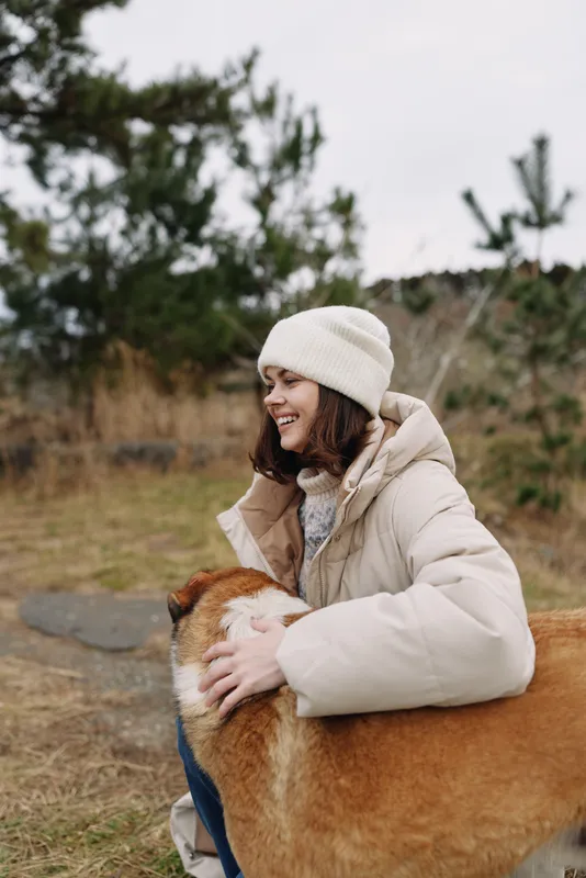 Woman, dog, outdoor, hug moment shows a woman warmly embracing a dog outdoors, smiling beside trees and grass, capturing cozy companionship and seasonal warmth in a quiet setting