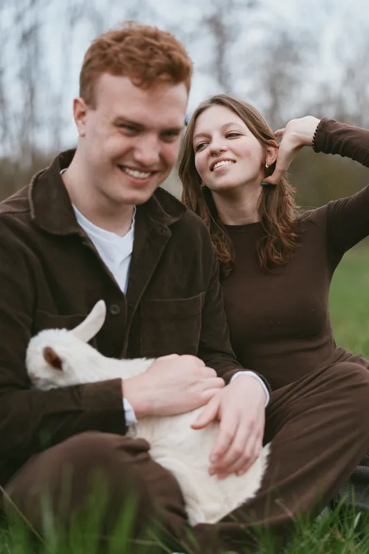 Young couple sitting on grass outdoors with a white baby goat, enjoying a relaxed moment in casual clothes during a lifestyle photoshoot in nature.