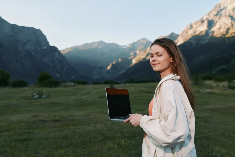 A young woman stands in a mountainous landscape holding a laptop, smiling softly as she works outdoors, blending nature and technology in a peaceful, productive moment.