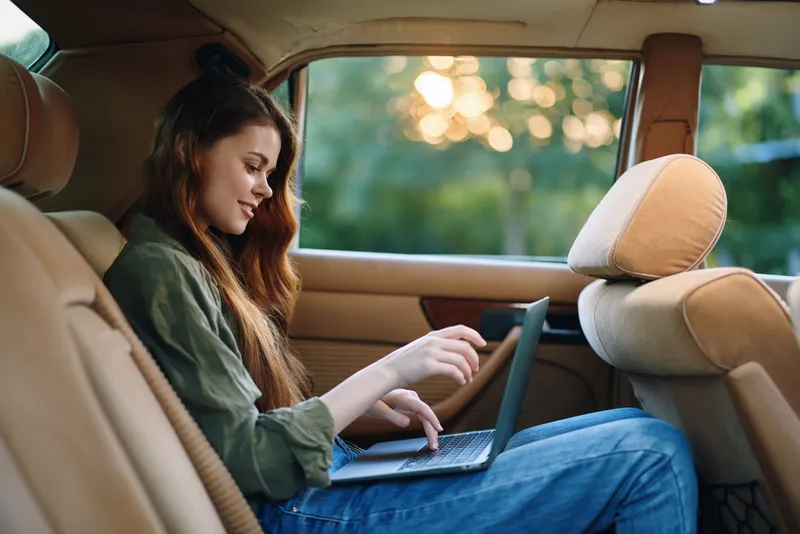 Young woman working on a laptop in the backseat of a car, dressed casually in a green shirt and blue jeans, enjoying a serene outdoor view, reflecting productivity and relaxation.