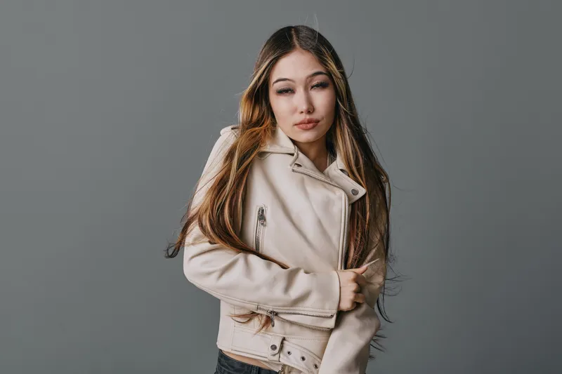 Young woman with long hair wearing stylish beige jacket posing with eyes closed on gray background in studio for fashion, portrait and beauty concept with casual look.