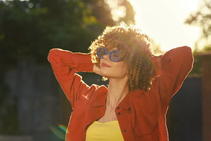 A stylish young woman outdoors in bright sunlight wears a red jacket and yellow top, standing with hands behind her head and a confident, relaxed expression