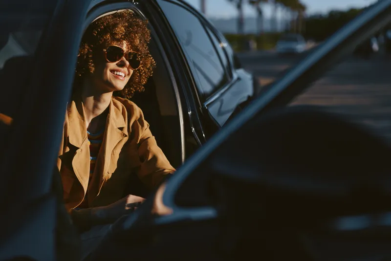 Woman in a vintage car, sunlit smile, retro film color, casual jacket, sunglasses, leaning out the window, candid lifestyle moment, warm natural light, relaxed outdoors vibe