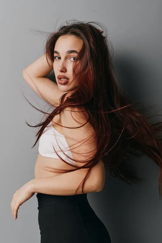 Young woman with long hair posing in studio, wearing white top and black pants, looking at camera with confident expression on gray background. Fashion, beauty and portrait concept.