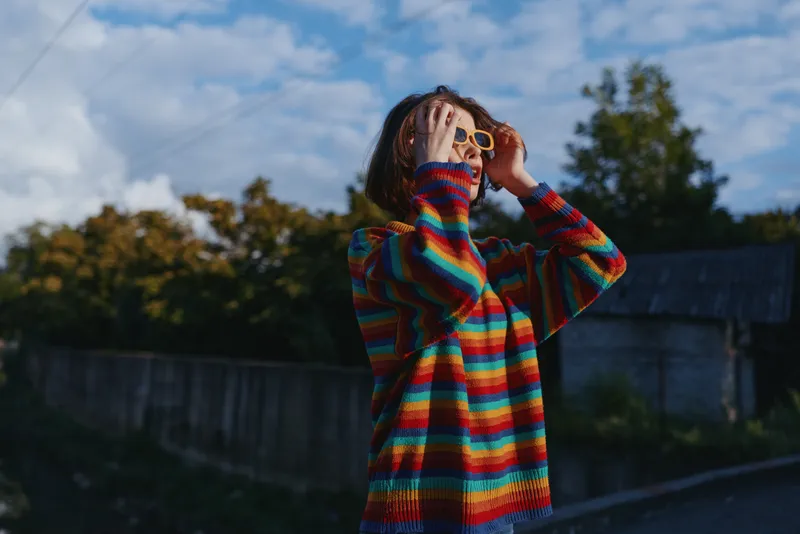 Woman in a colorful striped sweater and sunglasses outdoors, smiling with short hair in a casual outfit by a rural wooden fence, joyful portrait for lifestyle and fashion use.