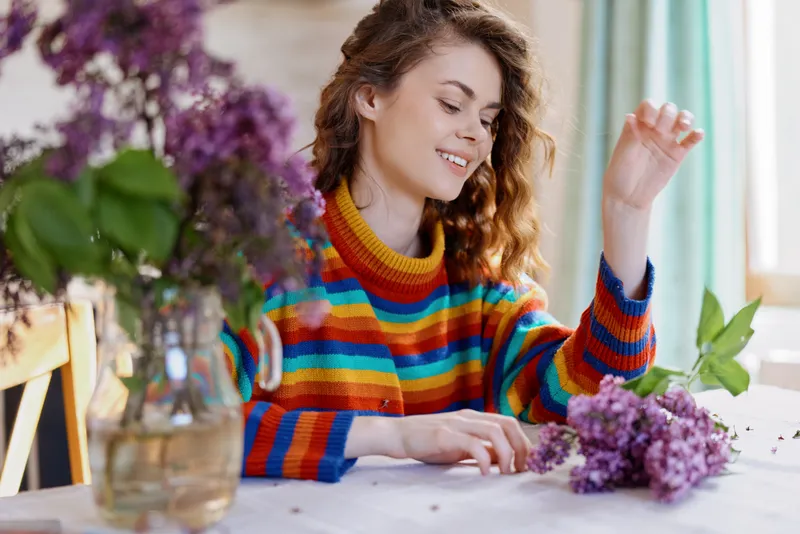 Cheerful woman indoors, arranging flowers at a sunlit table in a colorful striped sweater, capturing a relaxed lifestyle moment with natural colors, greenery, and cozy home ambiance