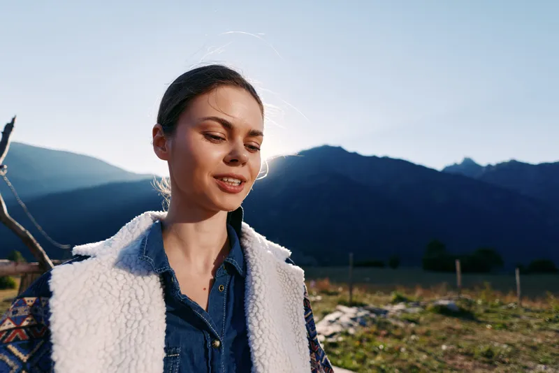 Woman portrait in mountains outdoors with warm smile and sunlight on her face, casual meadow outfit and denim vest, relaxed nature moment capturing a serene summer landscape.