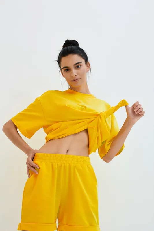 Young woman in bright yellow casual outfit tying shirt knot, posing with confident expression on plain white background, modern sporty style clothing.