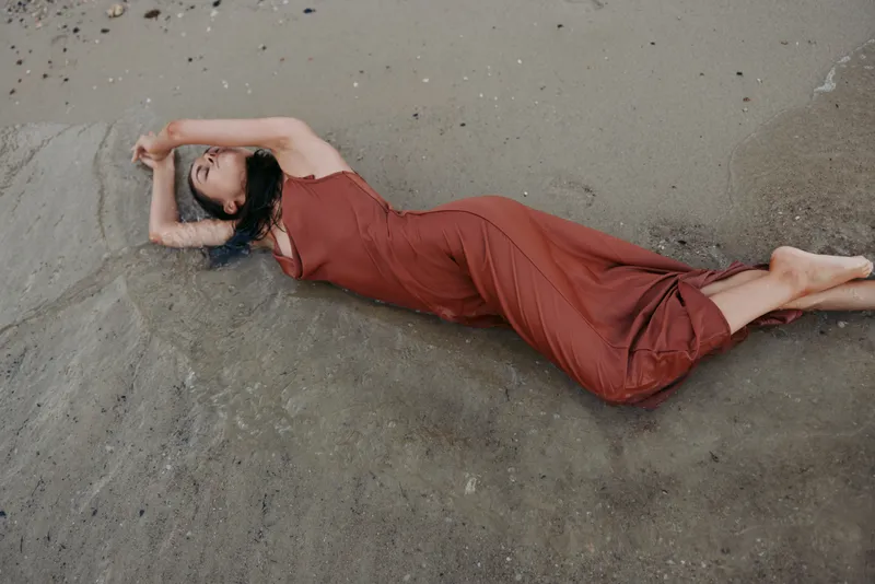 A solitary figure lies on a sandy shore wearing a flowing red dress, arms arched above, creating graceful lines and a serene mood with soft, natural light.