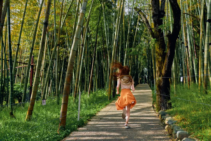 Girl running along a gravel path through a dense bamboo forest, wearing an orange skirt and backpack. Tall trees and dappled sunlight create motion and joyful travel in nature.