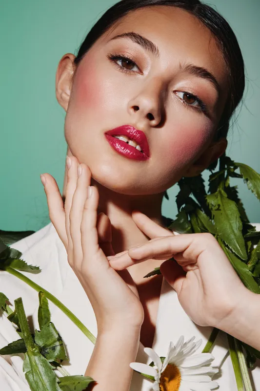 Close-up of a young woman with vibrant red glossy lips and flawless skin posing with hands near face surrounded by green leaves and white flowers. Beauty and fashion concept.