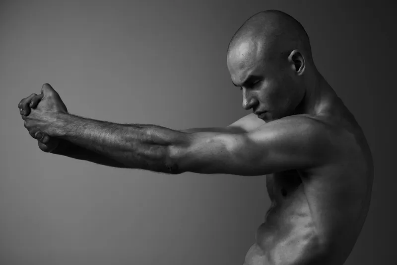 A focused man stretching his arms, showcasing strong musculature. The image is in black and white, highlighting the contrasts in body tones and shapes.