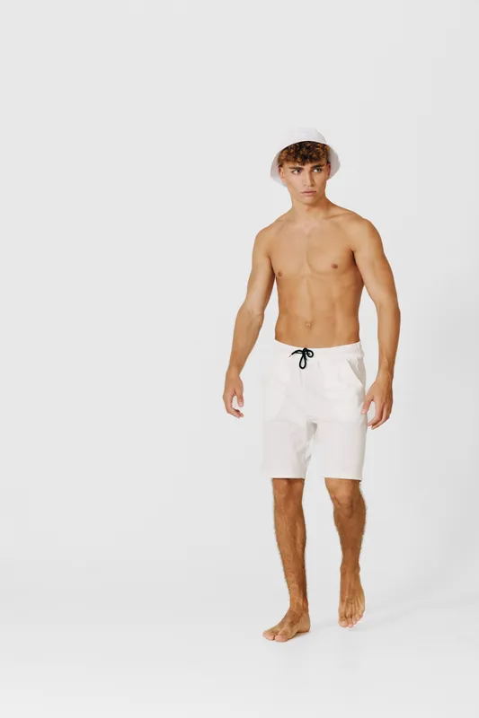 Young man with bare torso wearing white shorts and bucket hat walking barefoot on white background. Casual summer style with relaxed confident pose and minimal clothing.