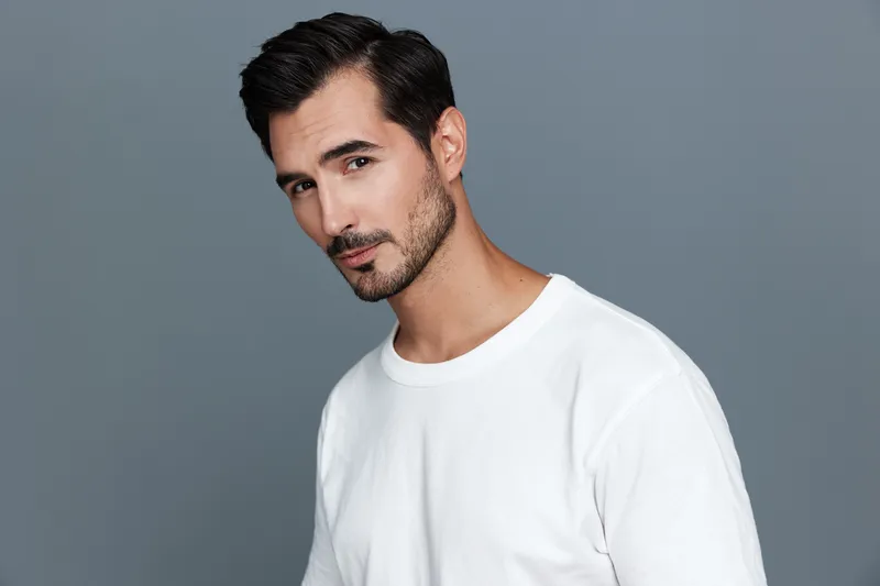 Handsome man with facial hair wearing a white t-shirt, looking directly at camera with a confident expression against a solid gray background. Portrait concept.