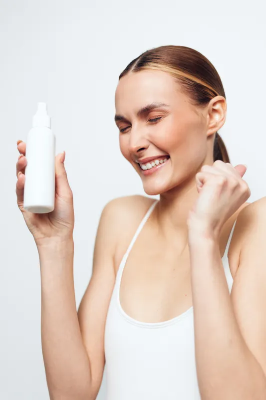 Happy young woman in white tank top holds white cosmetic bottle with closed eyes and smile, showing excitement and satisfaction with skincare product on white background.