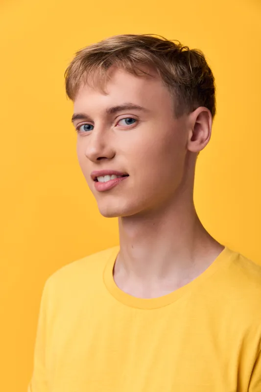 Adult male wearing a yellow shirt stands against a vibrant yellow backdrop, looking toward camera with a confident, friendly expression and relaxed posture for a bright studio vibe