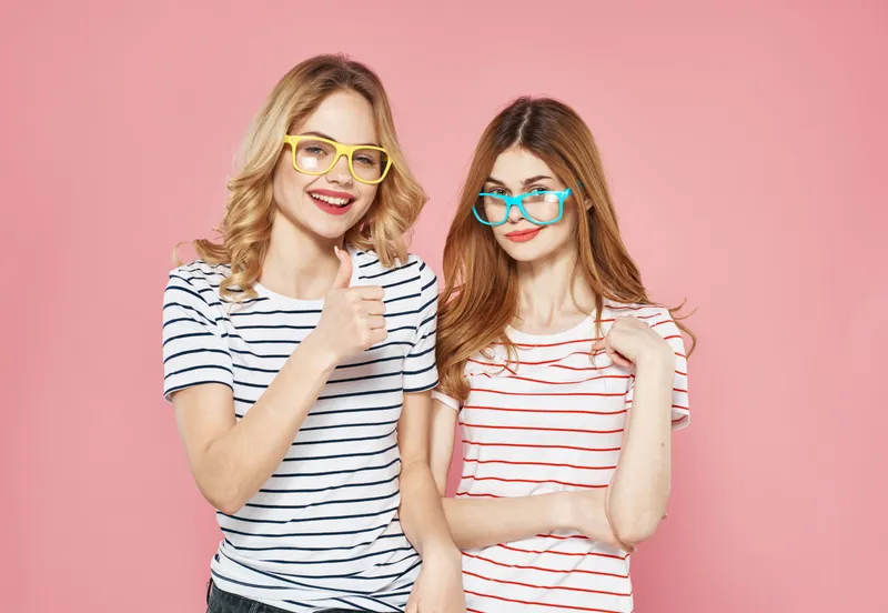 Две женщины в полосатых футболках и модных очках общаются, демонстрируя дружбу и единство на розовом фоне