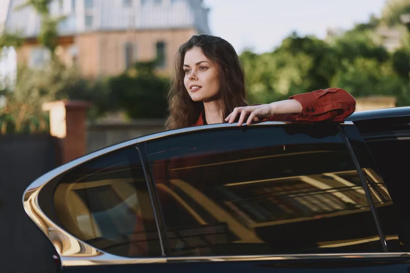 Smiling woman with curly hair leaning out of a car window, red blouse, outdoor city backdrop. Lifestyle moment emphasizing freedom, exploration, and casual confidence