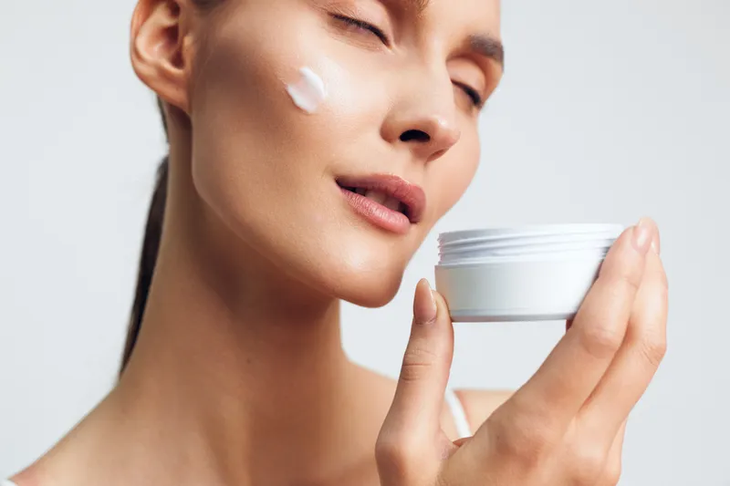 Closeup of woman applying face cream on cheek, eyes closed, smooth skin, natural beauty, soft light, skincare routine, white background, serene expression