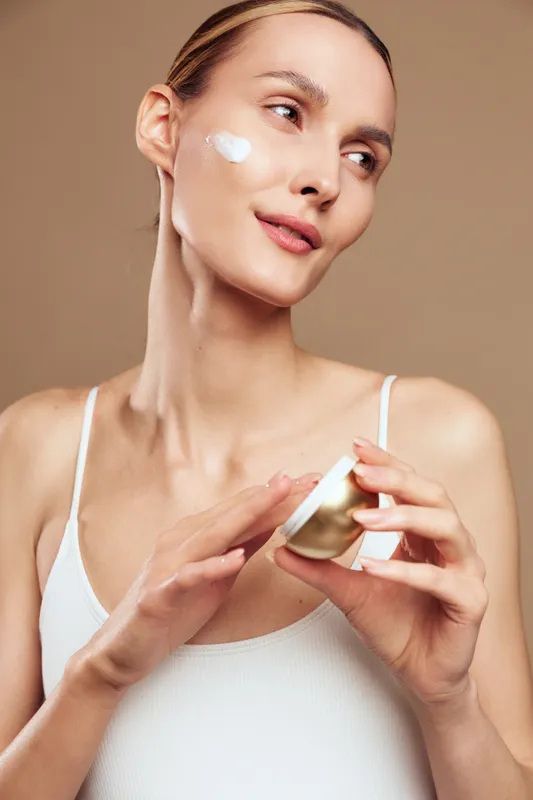 Young woman with clear skin applying white face cream on cheek. Close-up with beige background. Concept of skincare, beauty routine, self-care, and moisturizing treatment. Soft natural light.