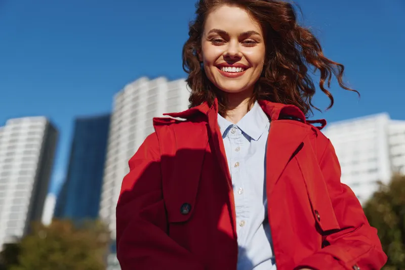Confident woman in a red coat smiles in an urban setting, bright day cityscape behind. A vibrant lifestyle portrait showcasing modern fashion and optimism.