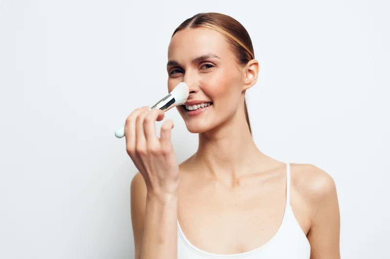 Young woman applying makeup with a brush smiling gently white background natural light closeup beauty routine skincare concept clean skin