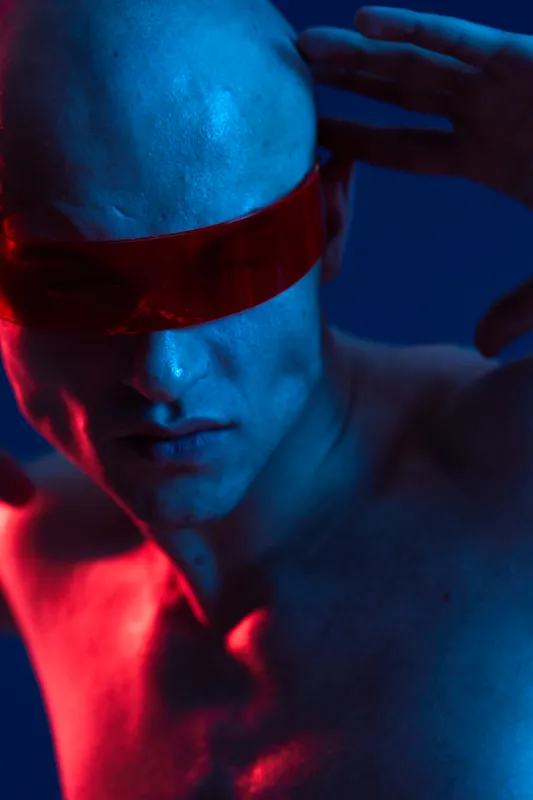 Man with striking expression wearing red sunglasses, showcasing an artistic blend of light and shadow in a bold blue environment. Black and white version available.