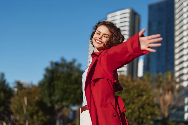 Cheerful woman outdoors smiling with arms outstretched in warm sunlight red coat city backdrop blue sky vibrant energy and carefree mood