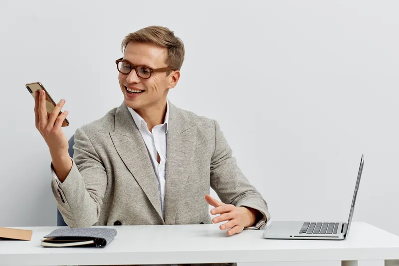 Smiling professional man in smart casual outfit holding a smartphone while working at a desk with a laptop and notepad, isolated on plain background. Business office lifestyle concept