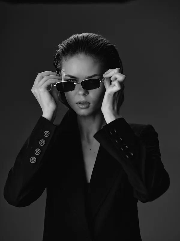 Black and white portrait of confident woman adjusting sunglasses in stylish dark blazer with serious expression in studio lighting.